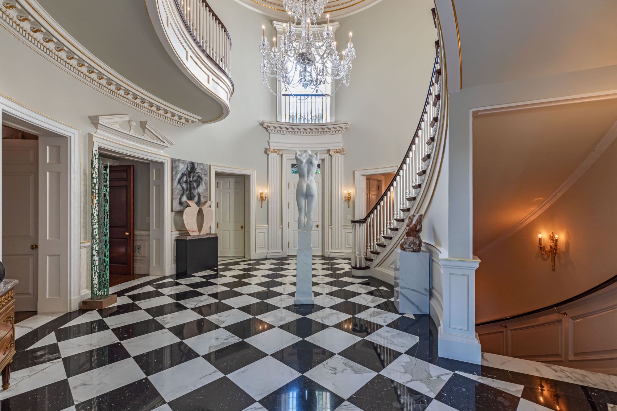 Elegant foyer with a grand staircase, luxurious chandelier, and striking sculptures against a checkerboard floor.