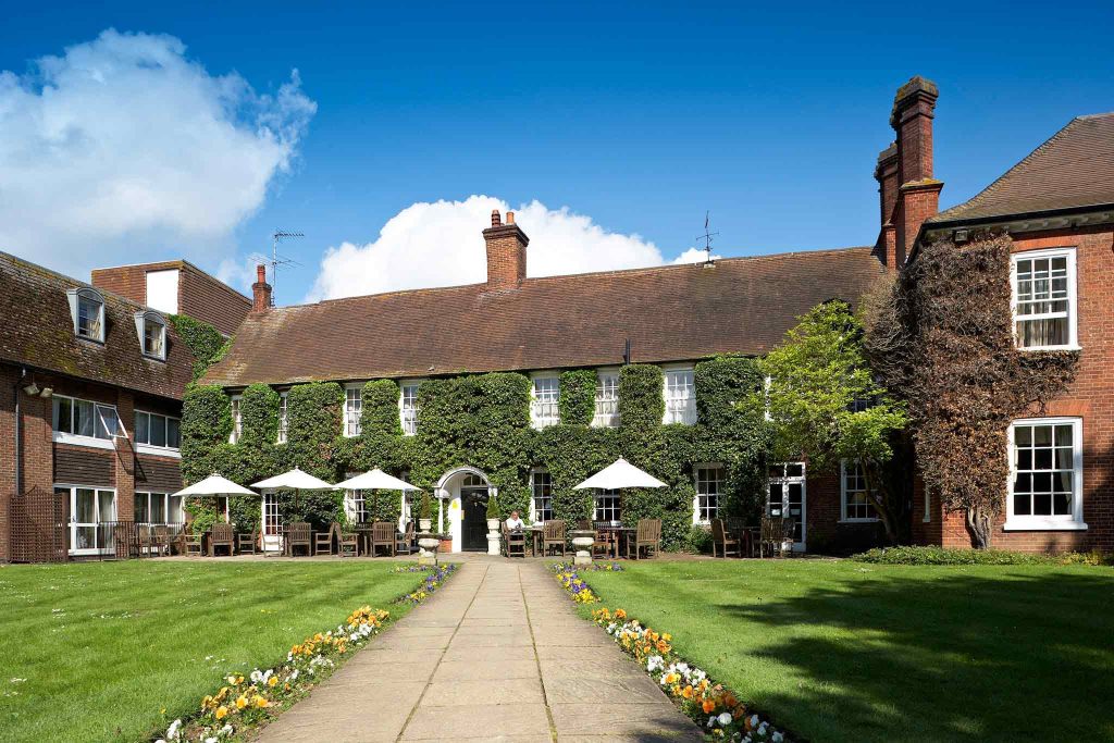 Historic grand inn in Surrey with lush ivy, outdoor seating, and a pathway bordered by colorful flowers under a blue sky.