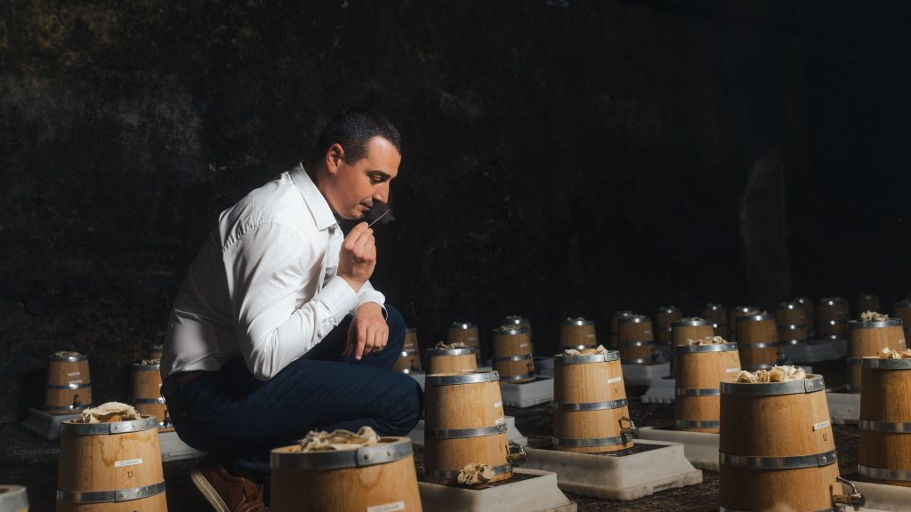 A person kneels near wooden barrels, thoughtfully inspecting notes in a dimly lit cellar, reflecting on cognac heritage.