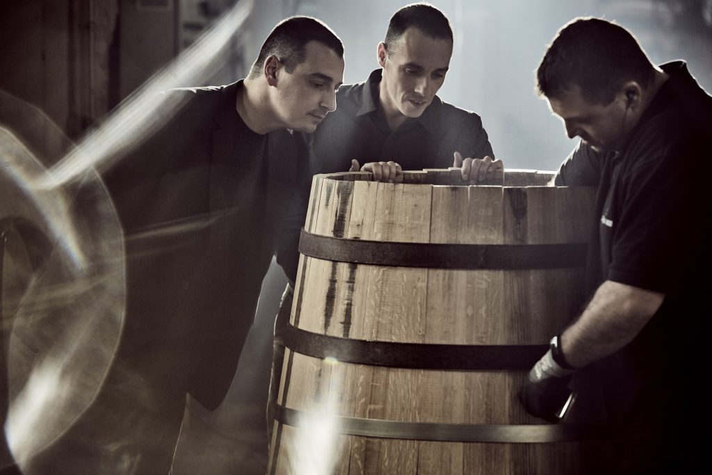 Three men inspect a wooden barrel in a dimly lit environment, emphasizing craftsmanship in cognac production.