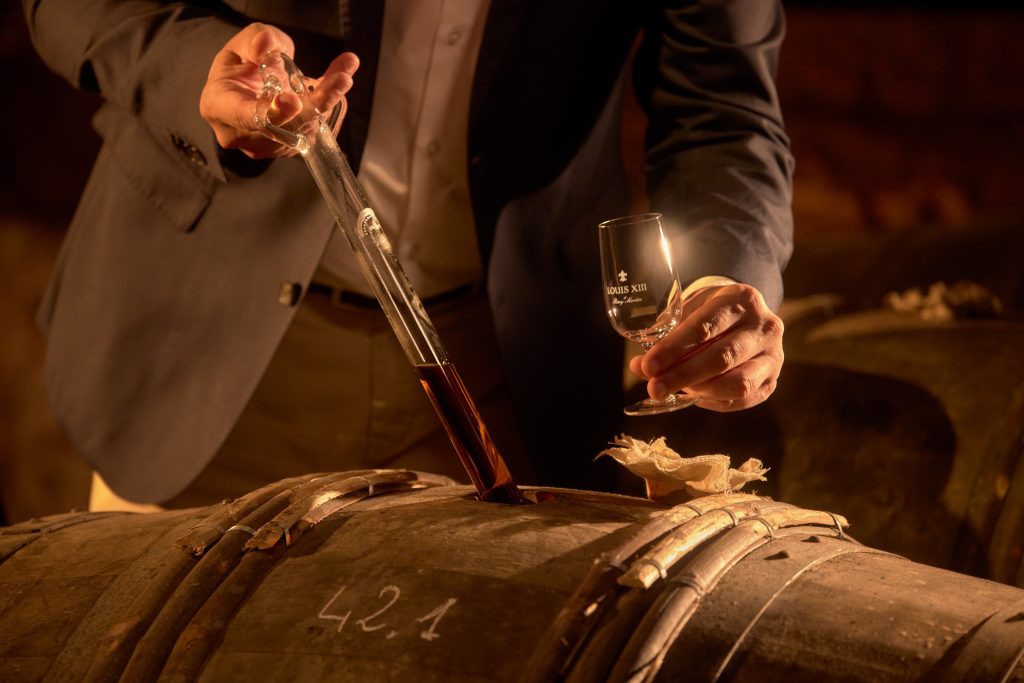 A person pours cognac from a tube into a glass, standing over a wooden barrel marked with the number 421.